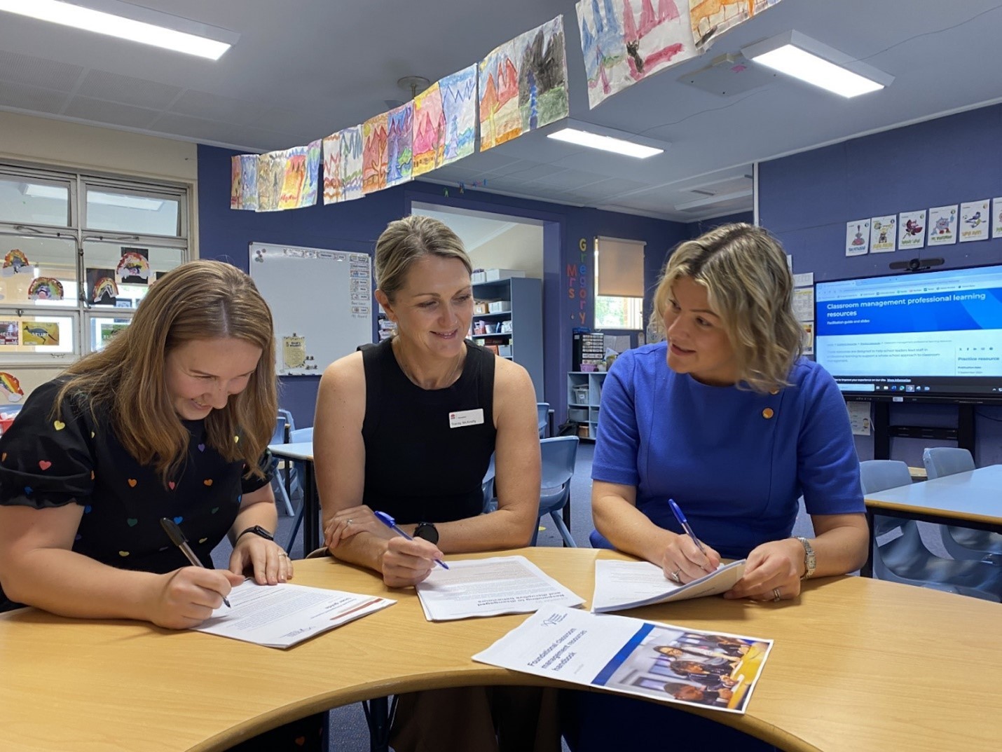 Three women sitting at a table in a classroom using and discussing AERO's classroom management resources.