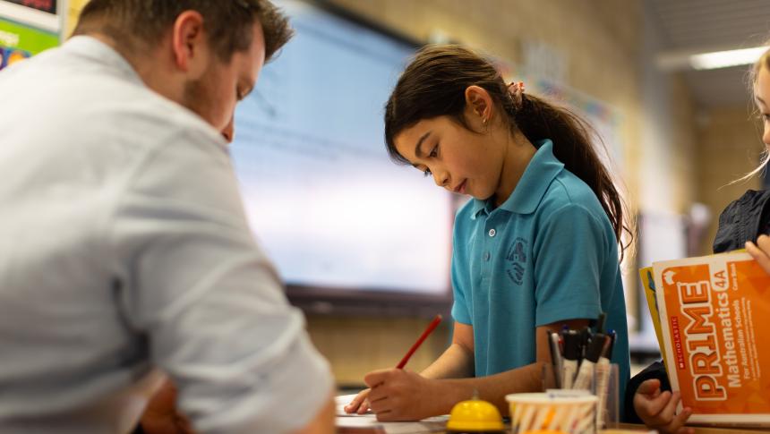 Primary school student standing in front of desk writing with pencil. Teacher sitting at desk helping student with writing.