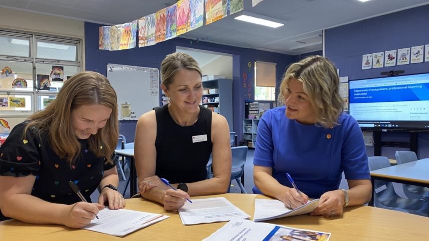 Three women sitting at a table in a classroom using and discussing AERO's classroom management resources.