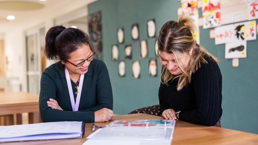 High school student sitting at desk with teacher discussing work.