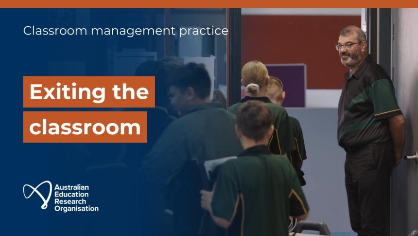 A teacher stands in the doorway of a classroom, smiling at students as they line up to exit.