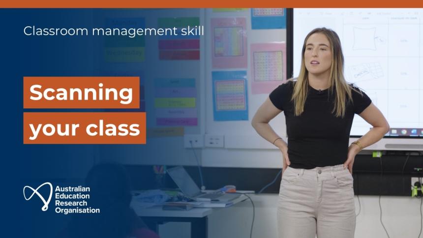 A screenshot from the video showing a teacher standing at the front of the class with hands on hips and gazing out across the classroom.