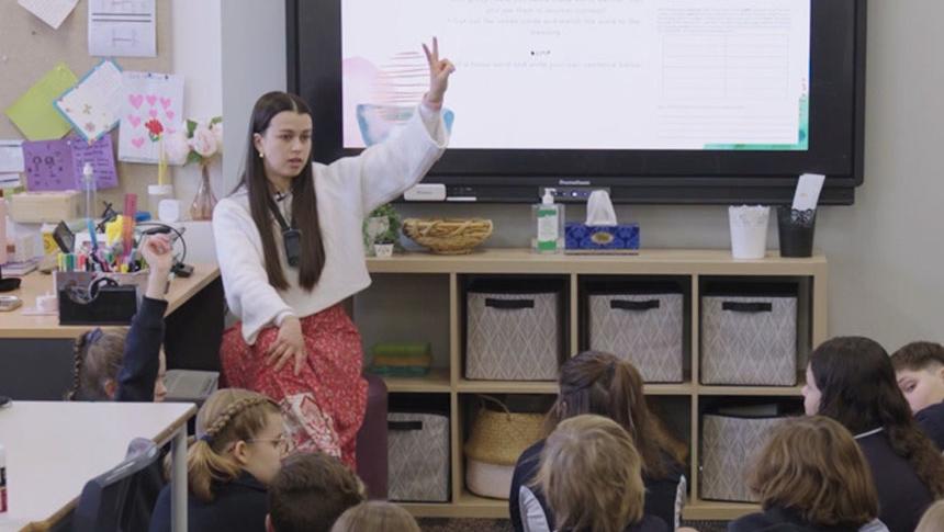 Teacher sitting on stool in front of students on floor holding up 2 fingers