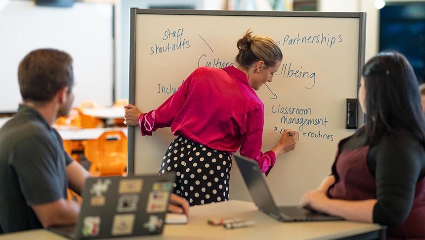 Teacher writing on whiteboard while 2 colleagues listen and take notes on laptops