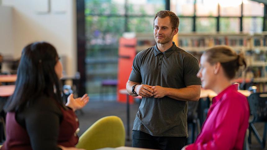 Teacher standing in front of 2 colleagues sitting at a table in library talking