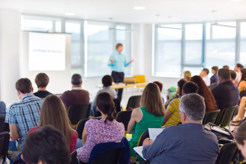 A lecturer stands in front of a room full of people sitting and watching them