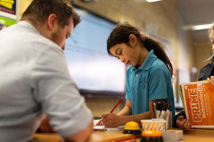 Primary school student standing in front of desk writing with pencil. Teacher sitting at desk helping student with writing.